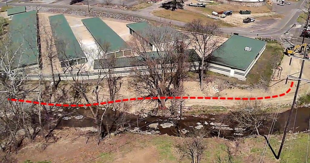 Aerial view showing a green-roofed industrial complex near a small stream. A red dashed line runs horizontally across the image, indicating a potential pathway or boundary parallel to the stream. Trees and a construction vehicle are visible.