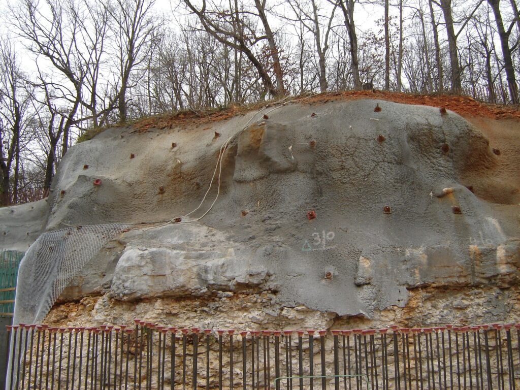 Rocky hillside with netting and anchors for stabilization. The slope shows exposed rock layers, with some concrete patches and a written "Δ 39°" mark. Leafless trees stand atop the hill, and metal fencing is visible in the foreground.