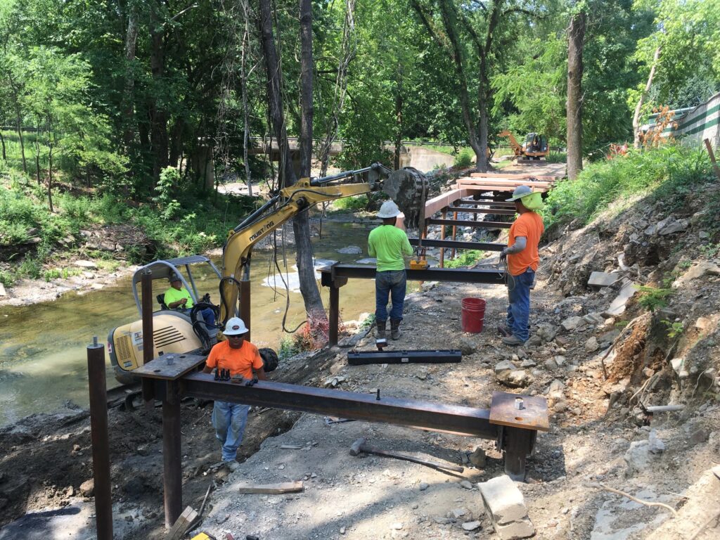 Construction workers in hard hats and bright shirts install a metal framework along a riverbank in a wooded area. A small excavator operates nearby, moving earth and materials for the project.