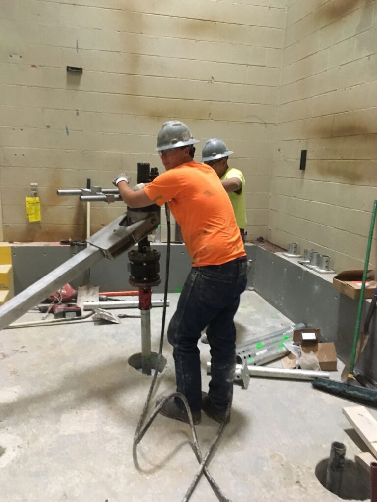 Two construction workers in hard hats and safety vests operate machinery indoors. One wears an orange shirt, while the other has a yellow one. The room has concrete walls and floor, with tools and materials scattered around.