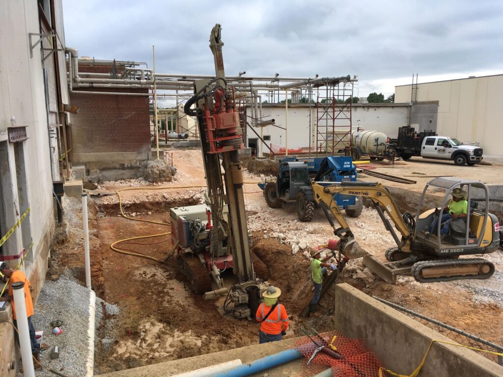 Construction site with three workers operating heavy machinery. A large drill and excavator are in use, surrounded by pipes and building materials. Overcast sky and industrial buildings in the background. Safety gear is worn by workers.