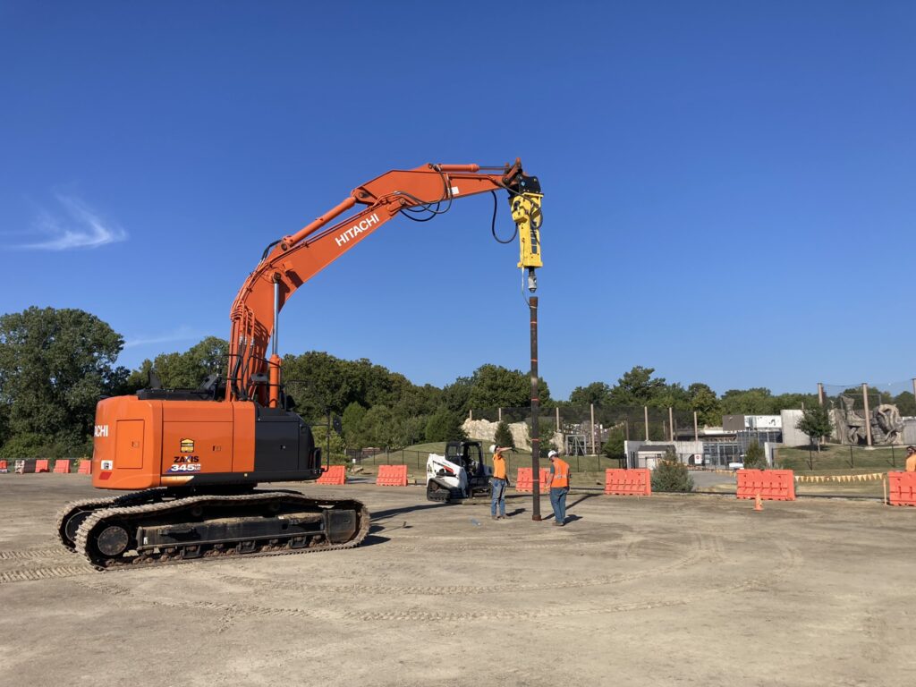 An orange excavator with a hydraulic breaker attachment is working in a construction site. Three workers in safety gear stand nearby. The site is surrounded by trees, orange barriers, and clear blue sky above.