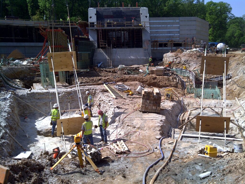 Construction site with several workers wearing safety gear, surrounded by excavation equipment, building materials, and structures under construction. A partially built concrete building is visible in the background, with scaffolding and tools around.