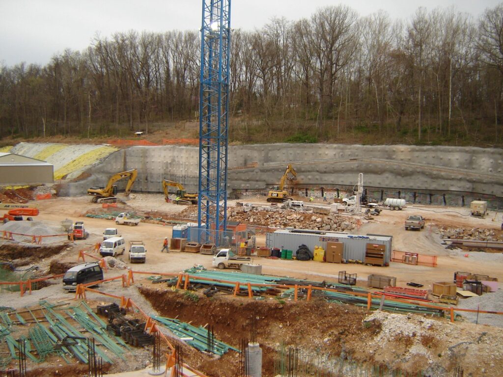 Construction site with several vehicles and machinery, including excavators and trucks, surrounded by trees. A tall blue crane stands at the center. Building materials are scattered around, including pipes and beams.