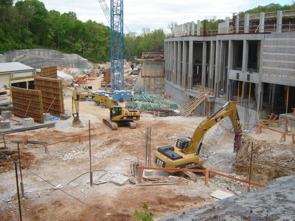 Construction site with two large excavators working on a dirt area. A multi-story building framework is visible on the right, while various construction materials and equipment are scattered around. A crane and dense green trees are in the background.