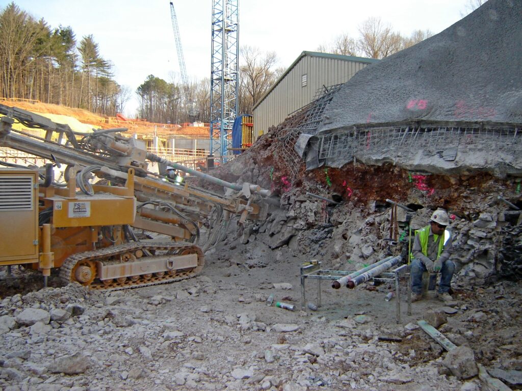 A construction site with heavy machinery digging into a rocky hillside. A worker in a safety vest and helmet is sitting on a metal structure nearby, with various construction materials scattered around. Cranes and trees are in the background.