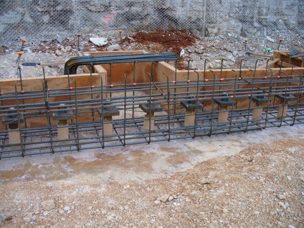 A construction site with a metal rebar framework and wooden forms set up for concrete pouring. The ground is uneven with dirt and rubble, and there's a chain-link fence in the background.