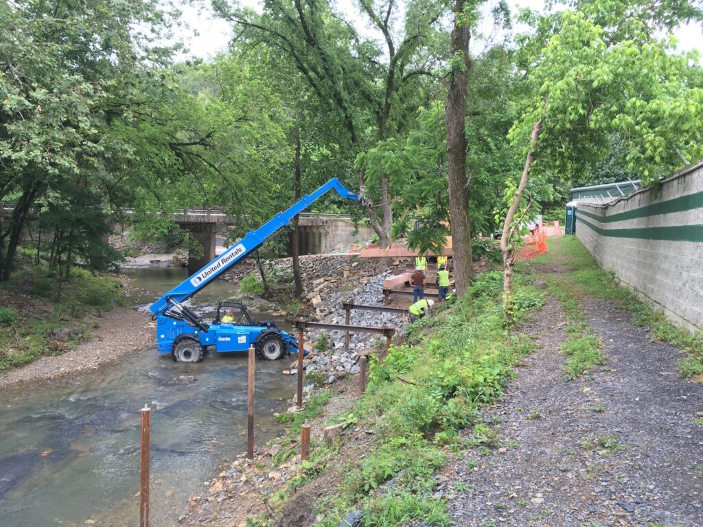 A blue crane is stationed by a creek, moving rocks near a wooded area. Workers in reflective vests are visible, working on the embankment. A gravel path runs alongside, with a concrete wall on the right. Trees and greenery surround the scene.