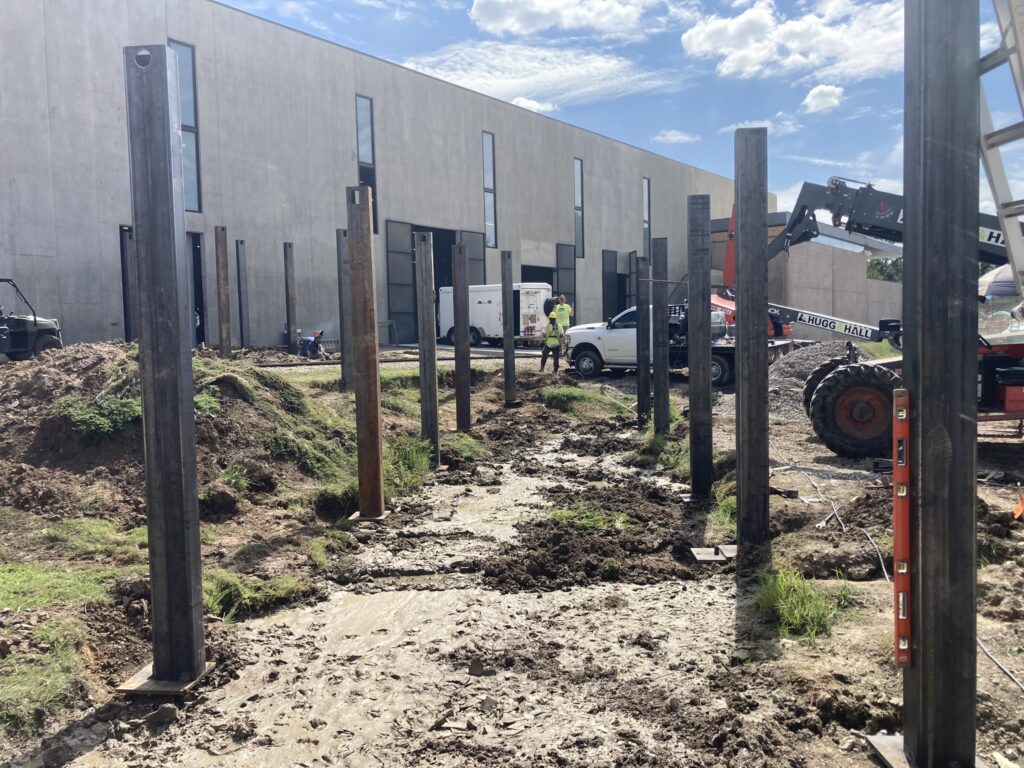 Construction site with metal beams in muddy ground, adjacent to a large industrial building. Workers and machinery are visible, with a blue sky overhead.