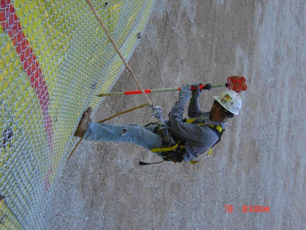 A construction worker in safety gear and helmet is secured with a harness while installing a piece of equipment on a chain-link fence on a concrete surface.