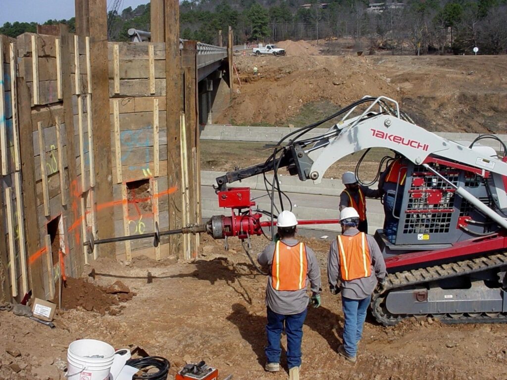 Construction workers in safety gear operate heavy machinery to bore into a wooden retaining wall near a bridge. The site is surrounded by dirt and construction materials, with trees and a road visible in the background.