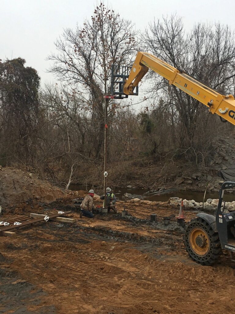 A construction site with two workers installing a pole using a yellow telescopic handler. The area is surrounded by bare trees and dirt. Equipment and construction materials are visible on the ground.