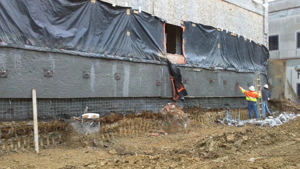 Two construction workers apply concrete to a foundation wall at a building site. The wall is covered with a black tarp, and the ground is muddy with scattered tools and debris. The workers wear safety gear and high-visibility clothing.