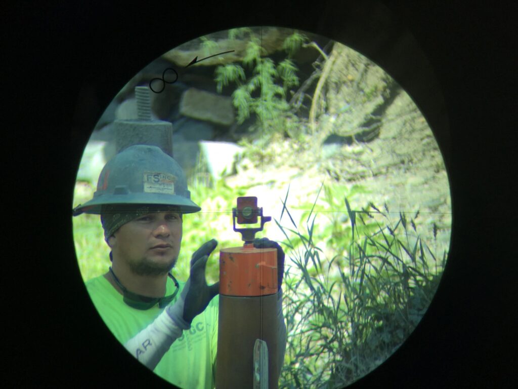 A construction worker wearing a hard hat and a green shirt is looking through a scope at a leveling rod in a grassy area. The scene is viewed through a circular lens, with a bolt visible on the left side.