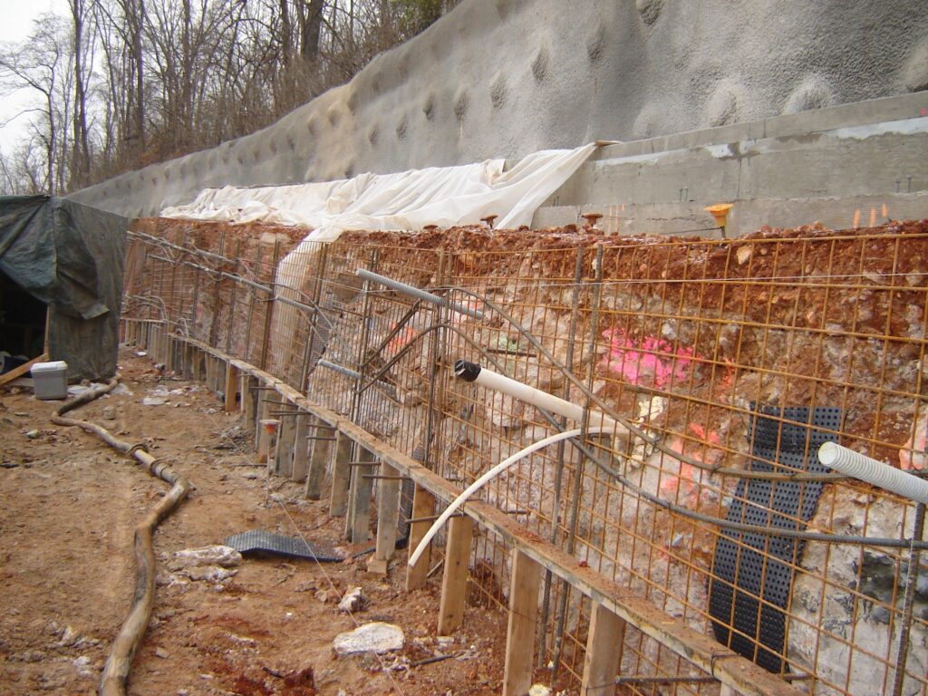 Construction site of a retaining wall with exposed rebar and drainage pipes. The wall is partially covered with white plastic sheeting, and the area is surrounded by loose soil and construction materials. Trees are visible in the background.