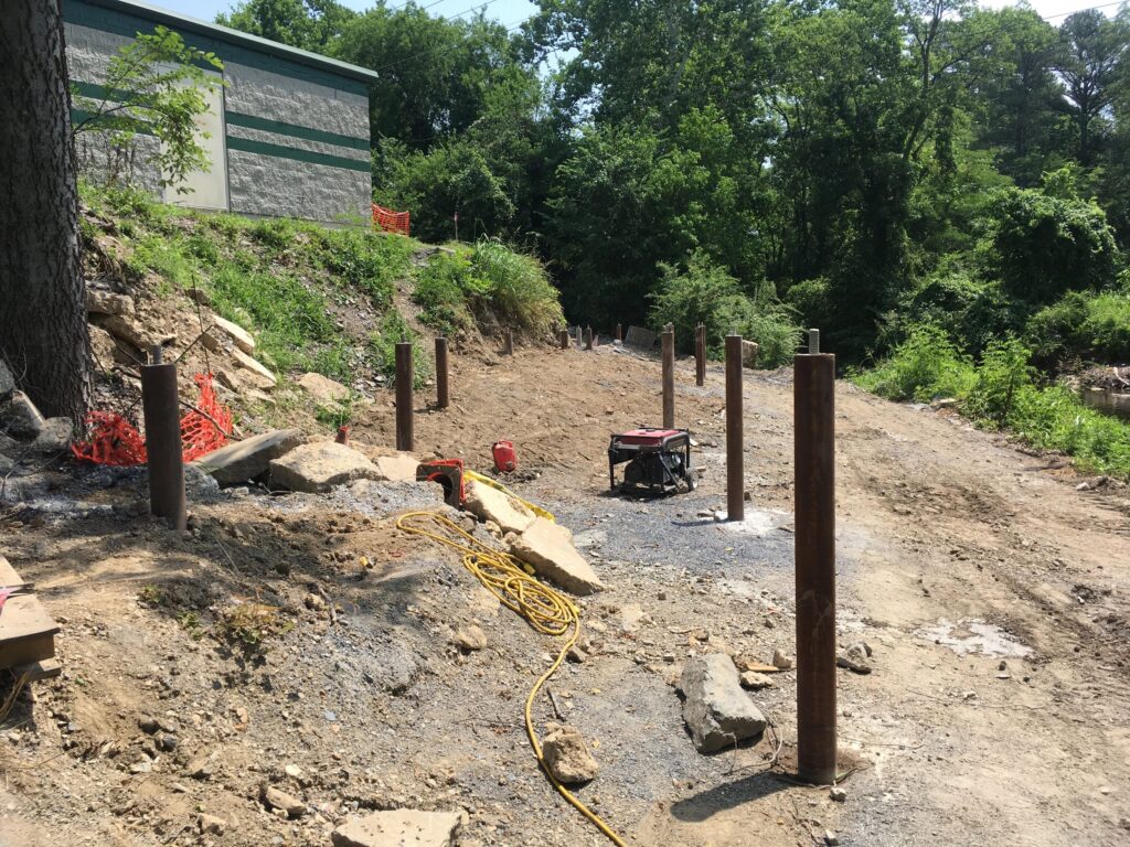 Construction site with several metal poles set into the ground along a dirt path. A few tools and cables are scattered nearby. A partially built structure is visible on the left, surrounded by trees and greenery.