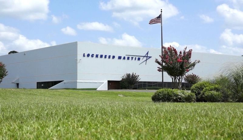 A large white building with the Lockheed Martin logo and name, surrounded by green grass and a few small trees. An American flag is flying on a pole in front of the building under a partly cloudy sky.