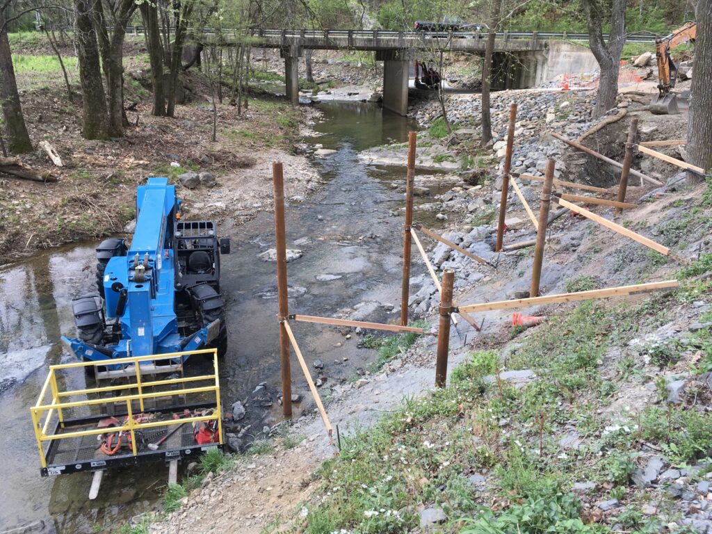 A blue construction vehicle is positioned on a riverbank near a shallow stream with a metal bridge. Wooden poles and beams are partially installed along the bank on uneven terrain. Trees and grassy areas surround the site.