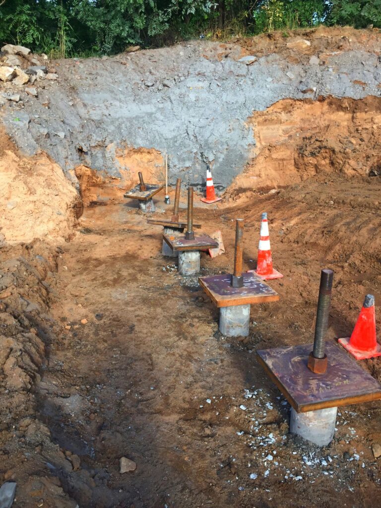 Construction site showing several concrete piles with metal rods protruding, evenly spaced in a trench. Orange traffic cones are placed near the structures. The background features a dirt slope and green foliage.