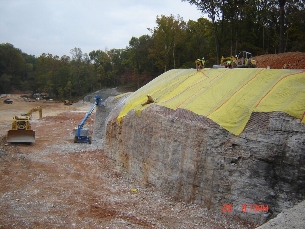 Construction site with workers securing a large yellow tarp over a rocky slope. Heavy machinery, including bulldozers and a cherry picker, are visible. The area is surrounded by trees and the ground is covered with gravel.