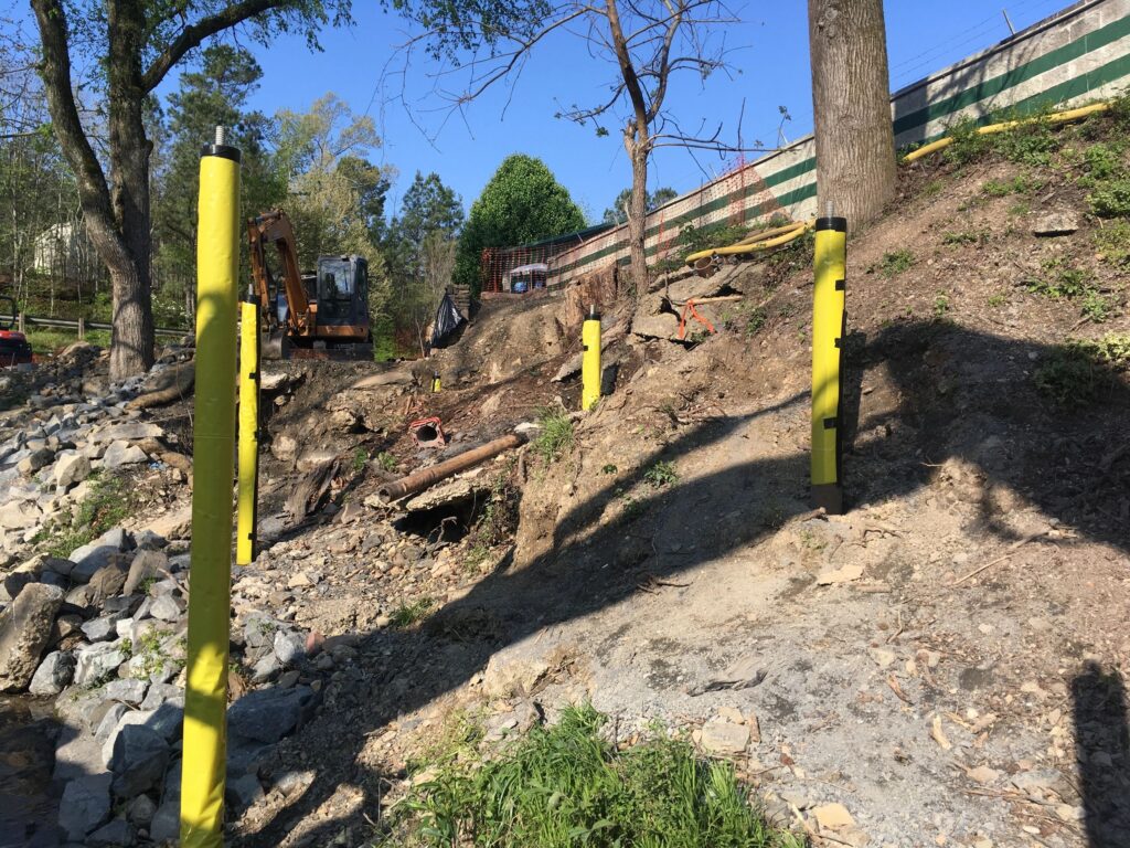 A sloped construction site with yellow poles, an excavator, and rocky terrain. Trees and a wire fence are visible in the background under a clear blue sky.