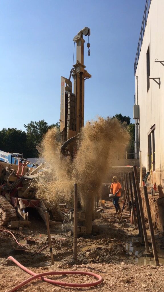 Construction site with a large machine drilling into the ground, causing a spray of dirt and debris. A construction worker in a helmet and orange vest stands nearby, surrounded by various equipment and tools.