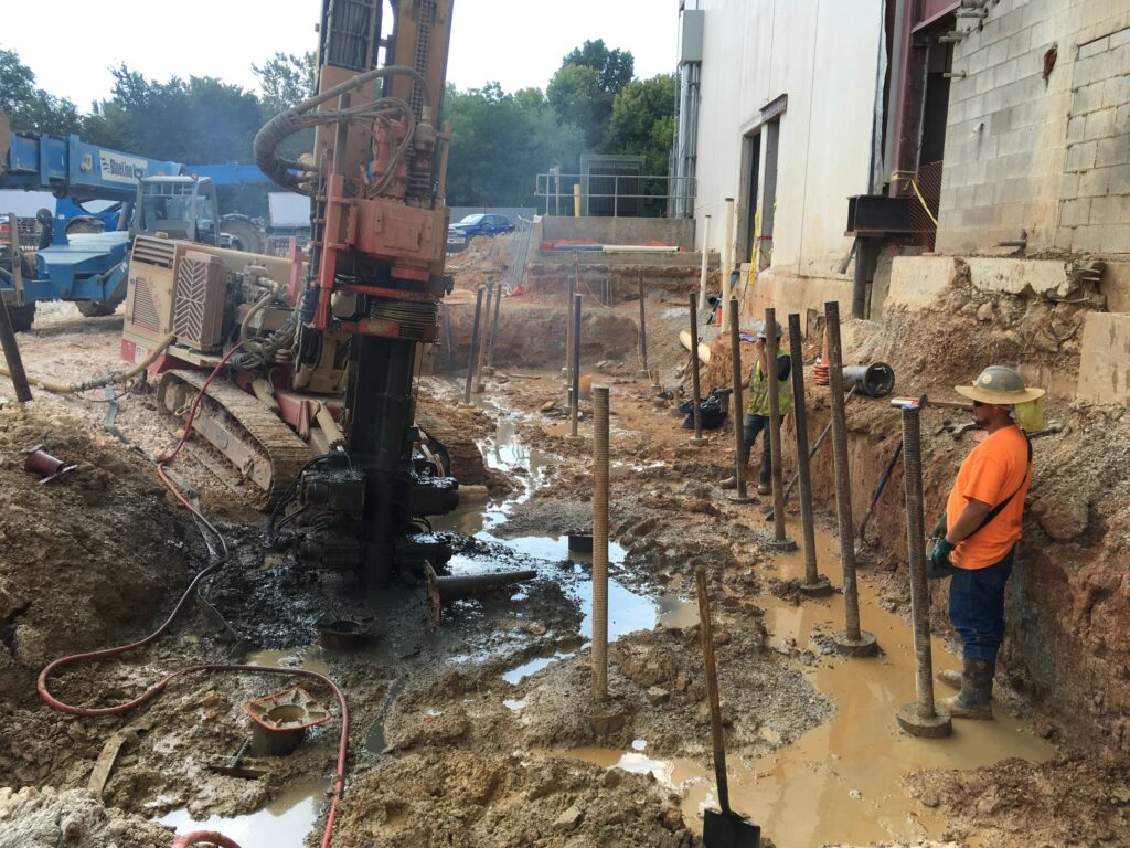 Construction site with workers wearing safety gear near a large drilling machine. The ground is muddy with several poles and pipes protruding. A partially constructed building is visible on the right, and machinery is in the background.