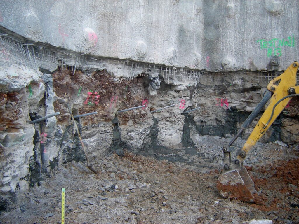 Construction site with an excavator arm digging into a layered rock and soil wall. Various rebar rods protrude from the wall, and numbers in pink chalk are marked across the surface. The ground is covered with loose dirt and debris.