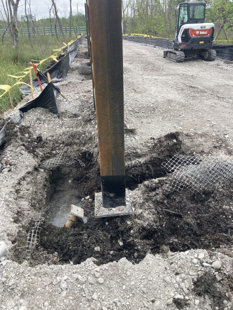 A construction site with a large vertical metal beam set in concrete. The ground around it is dug up, exposing soil and metal mesh. A small excavator is in the background, with trees and safety barriers nearby.