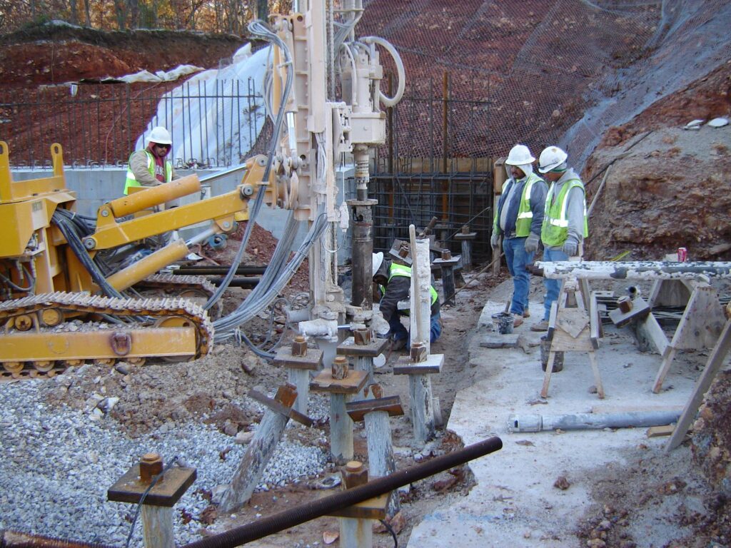 Construction workers in safety gear operate heavy machinery on a rocky site. They are drilling into the ground, surrounded by construction equipment, rebar, and steel mesh. The area is uneven and appears to be part of a larger building project.