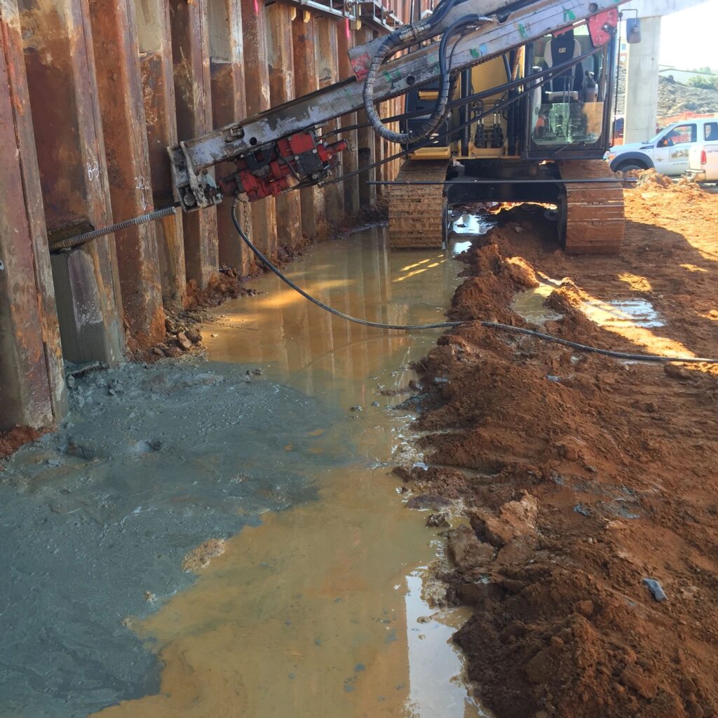 Construction site with a large drill rig working on a muddy area. Wet concrete is being poured into a trench. A parked car is visible in the background. Rusty steel sheet piles line the side of the site.