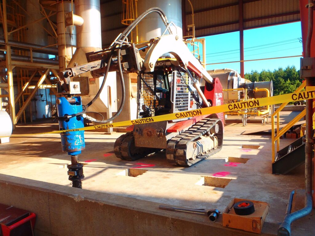 A small construction vehicle with a drilling attachment operates at a worksite, surrounded by caution tape. The site appears to have concrete with holes marked by pink paint. Metal structures and equipment are visible in the background.