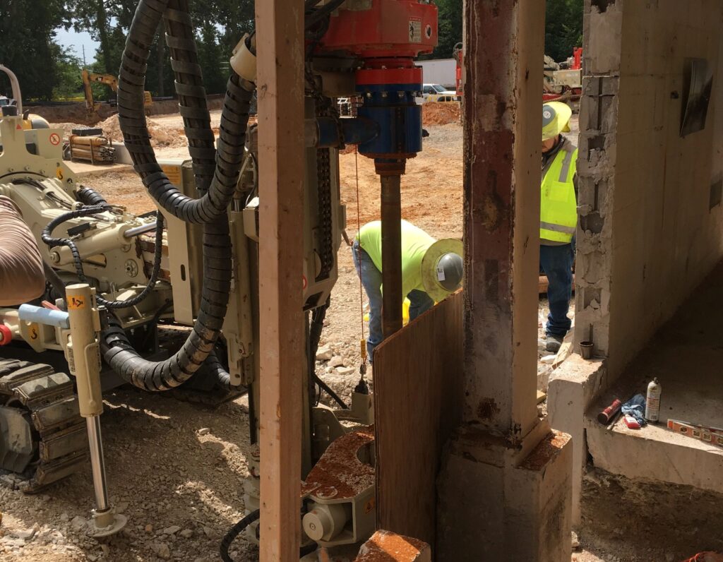 Construction workers using heavy machinery to drill into the ground at a construction site. They are wearing safety gear, including helmets and high-visibility vests, surrounded by dirt, machinery, and building structures.