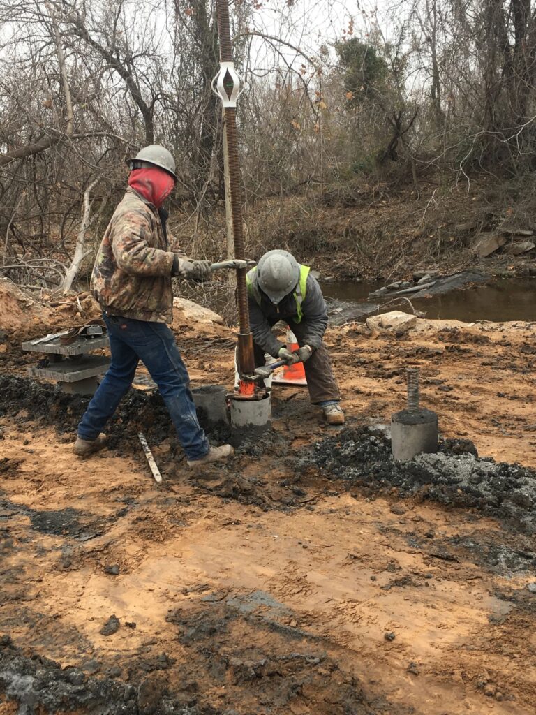 Two construction workers in safety gear are installing a utility pole in a muddy area near leafless trees and a stream. One is holding the pole steady while the other works at the base. The scene suggests a rural setting in autumn or winter.