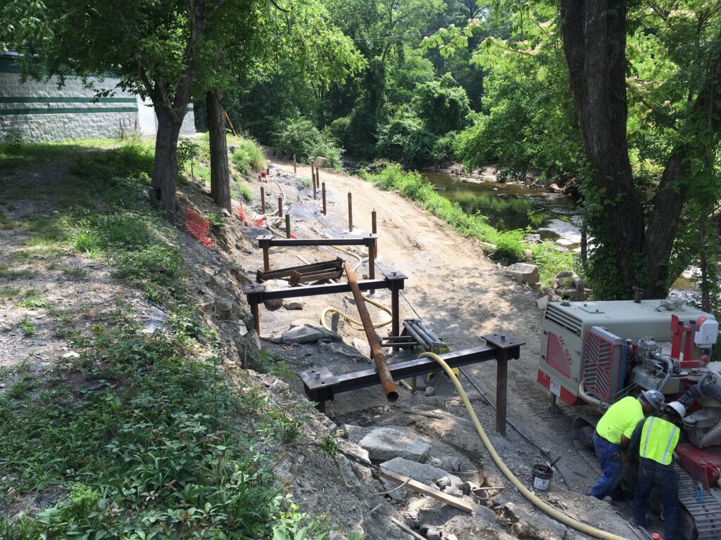 A construction site near a river with lush green trees. Workers operate machinery, and metal beams are positioned along a dirt path. Safety cones and orange netting mark the area, which is surrounded by dense vegetation.