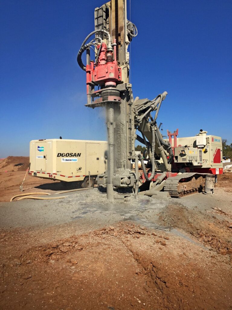 A large drilling machine is actively operating at a construction site, boring into the ground. Dust and debris surround the equipment. The sky is clear and blue, and the ground is covered with reddish-brown soil.