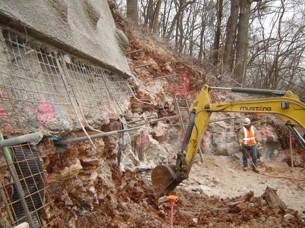 A construction worker in a safety vest and hard hat operates an excavator near a rocky hillside. The area shows exposed rock, rebar, and pink markings. Trees are visible in the background, suggesting a hillside stabilization project.