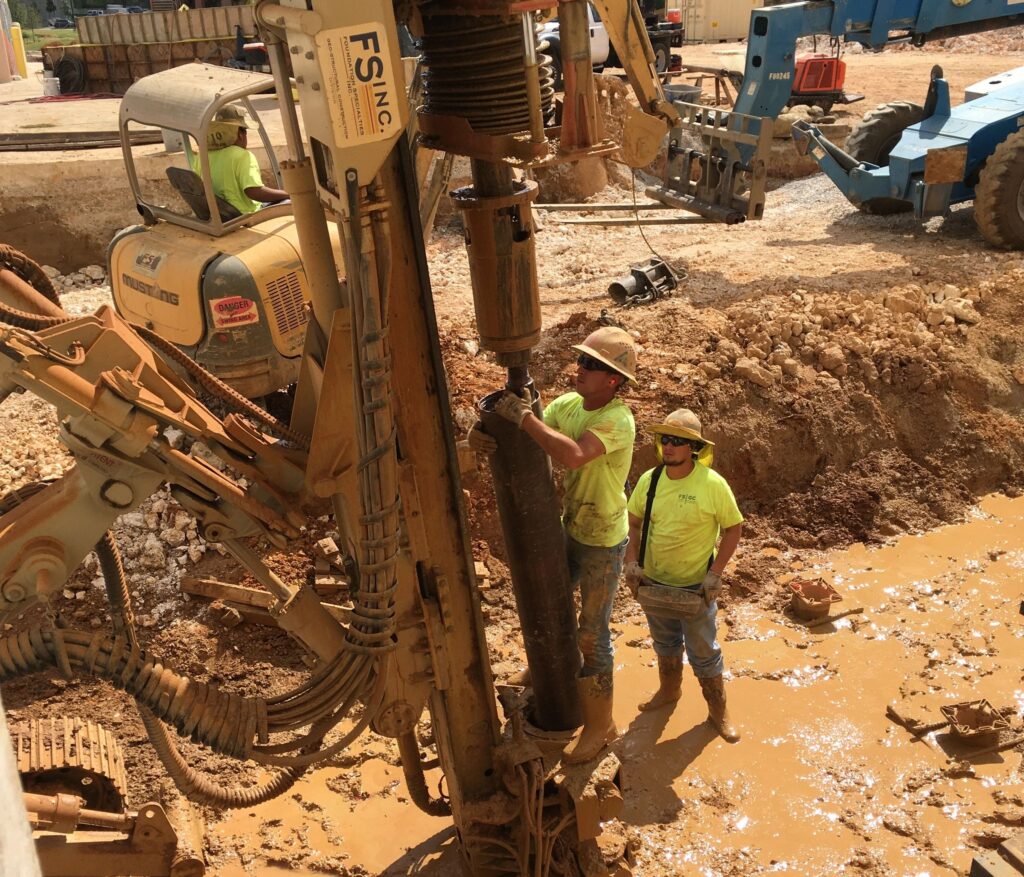 Construction workers operating heavy machinery at a muddy construction site. One worker is adjusting a large drill attached to an excavator while another observes. Various equipment and vehicles are visible in the background.