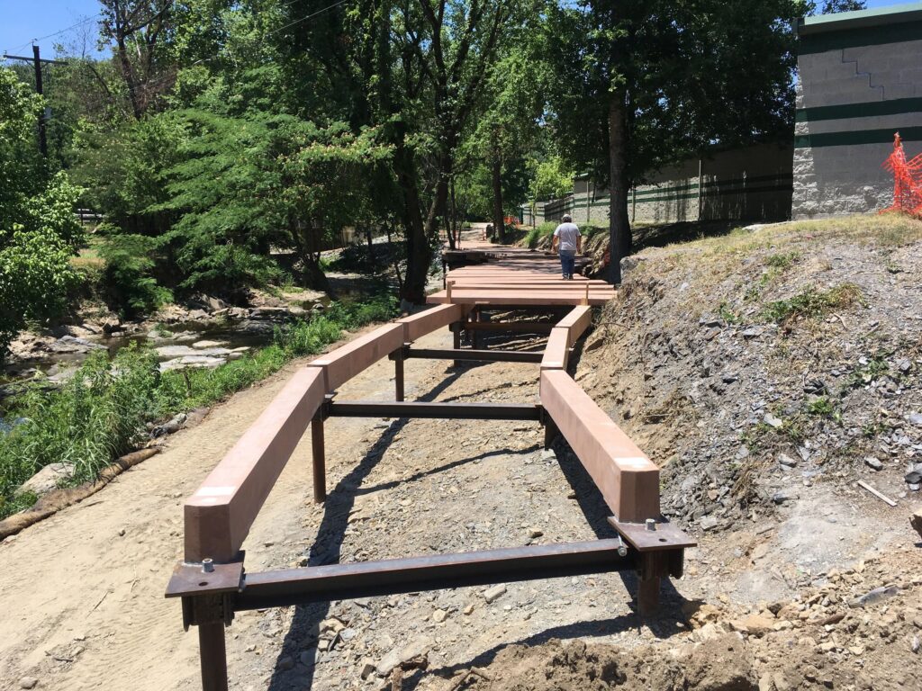 A construction worker stands on a partially built wooden bridge over a dry, rocky area with greenery. The bridge structure is supported by metal beams, and it's a sunny day with clear skies.