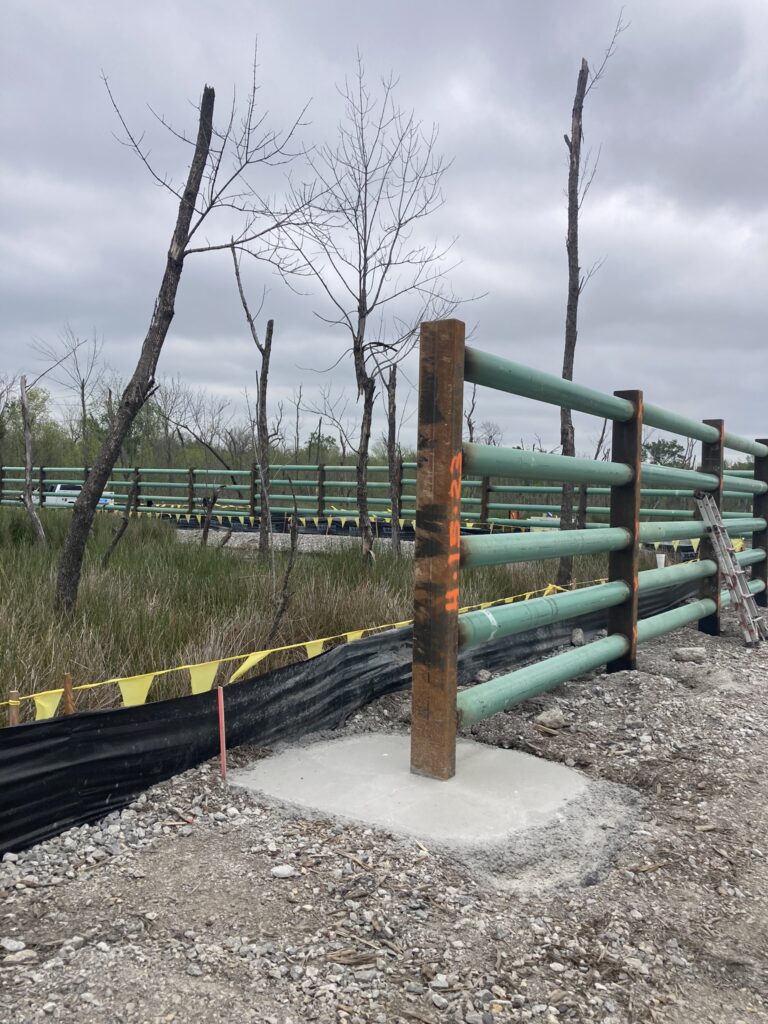 A construction site featuring a segment of a green metal fence with exposed beams and a concrete foundation. Yellow caution tape is visible alongside gravel and sparse, leafless trees under a cloudy sky.