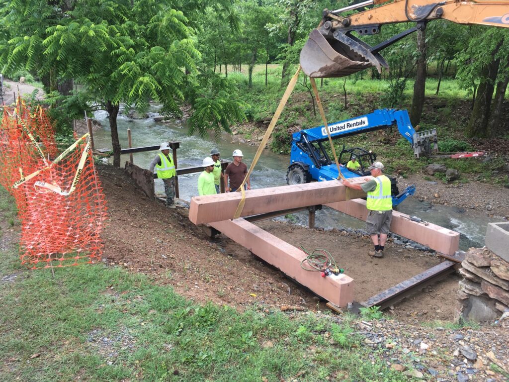 Workers in safety vests install large beams near a creek using construction equipment. Trees and a rural setting form the background, with orange safety fencing on the left. A blue lift vehicle is visible in the scene.