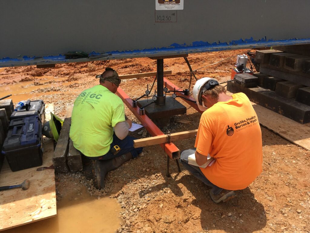 Two construction workers in safety helmets and high-visibility shirts install a support beam at a construction site. They are kneeling on the ground, surrounded by tools and materials, focused on securing the beam in place.