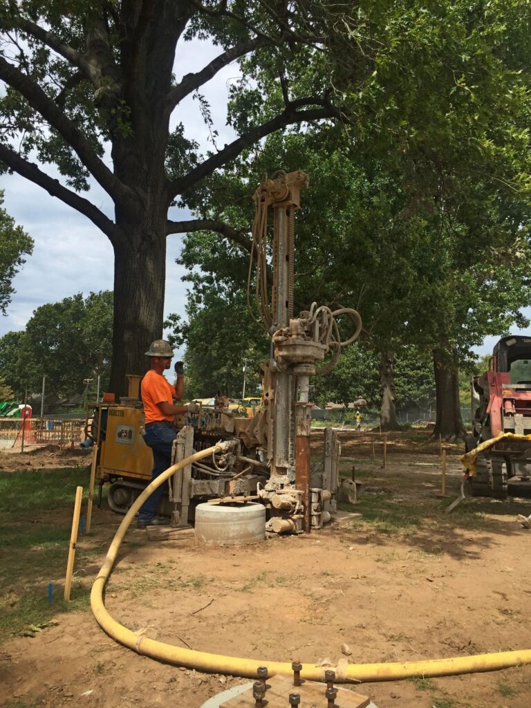 A worker in an orange shirt operates a large drilling machine in a wooded area. The scene is outdoors with green trees and a clear sky. Yellow hoses and equipment are visible on the ground.