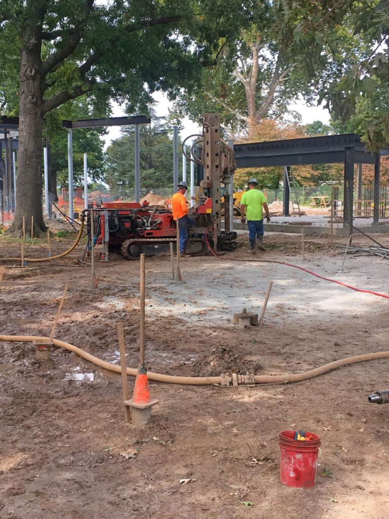 Construction site with workers in orange and green safety vests operating drilling equipment. The area is muddy, surrounded by trees and metal structures. A red bucket and orange cones are on the ground. The sky is partially visible through the trees.