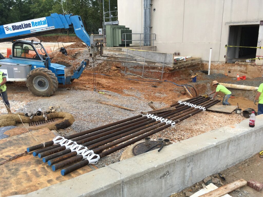 Construction workers are laying out long metal pipes on a gravel area at a construction site. A blue telescopic handler is nearby, with a partially constructed building in the background. Workers are wearing safety gear and hard hats.