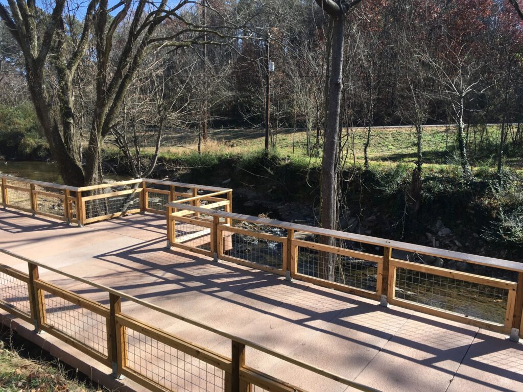 A wooden bridge with wire railings spans over a small creek. Sunlight casts shadows on the bridge's surface. Leafless trees and sparse grass line the banks of the creek on a clear day.