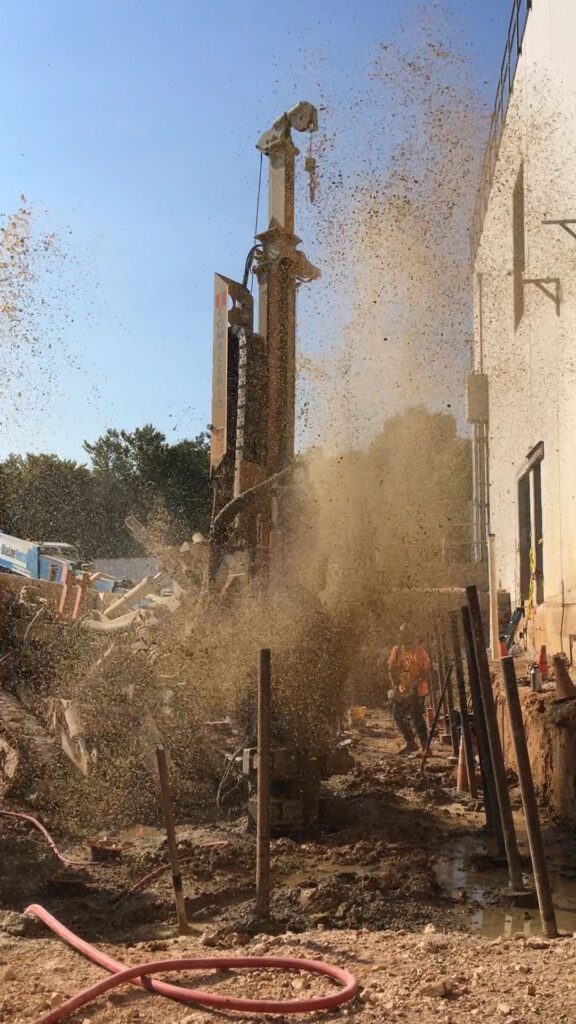 A construction site with a large drill releasing a spray of debris and dirt. Workers, wearing safety gear, manage the equipment. The scene is outdoors on a sunny day, with nearby buildings partially visible.