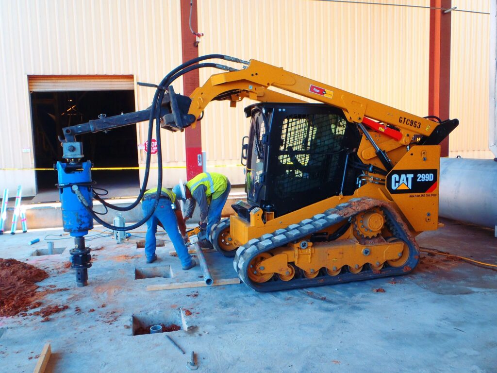 A construction worker operates a yellow CAT 299D compact track loader with an auger attachment. The vehicle is drilling a hole in a concrete floor inside a partially constructed building.