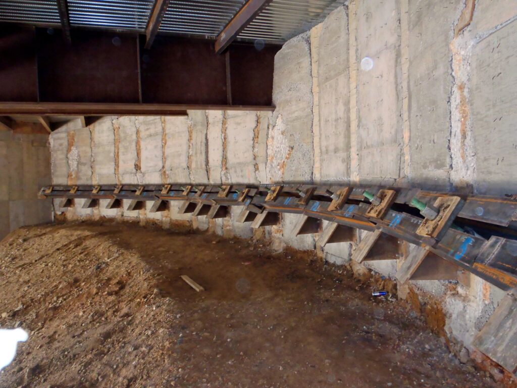 Interior construction site with exposed concrete walls and a curved metal framework partially embedded in the ground. A dirt floor covers the area, and overhead, a corrugated metal roof is visible.