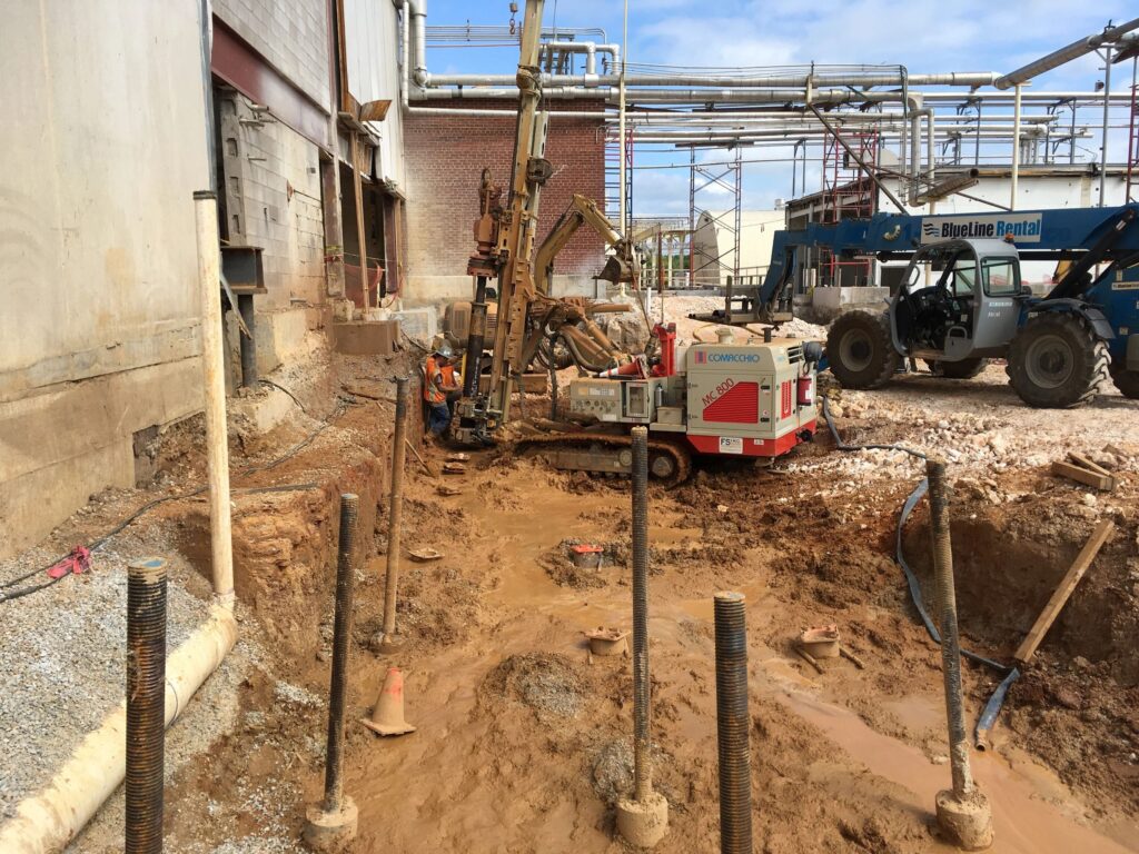 Construction site with workers operating drilling equipment next to a building. A crane and various materials are visible. The ground appears muddy, and several metal rods are installed in the earth. Scaffolding and pipelines are overhead.